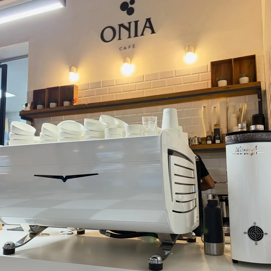 La cafetera blanca y el molinillo bajo el logo de Onia, con tazas apiladas encima
